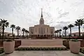 A landscape picture of the Red Cliffs Utah temple, with a sign saying it belongs to the Church of Jesus Christ of Latter-day Saints. Palm trees are seen on the side leading to the center.