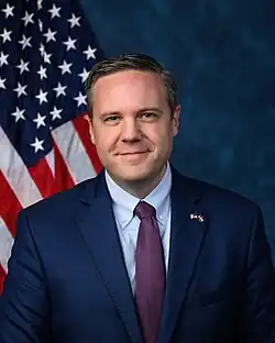 Official House portrait of Hurd in front of the U.S. flag, wearing a dark blue suit with American and Colorado flags lapel pin, light blue shirt, and checkered red and blue tie.
