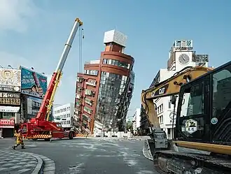 A large building at center leans forward. Heavy machinery is visible in the midground and foreground. A man walks at left.