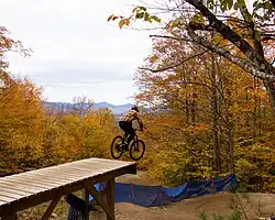 A mountain biker hits a wooden jump on Maine Line, Mt. Abram’s advanced downhill trail known for mandatory drops and gap jumps. Bright fall foliage fills the background.
