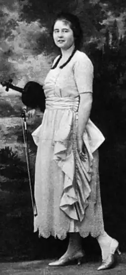A young white woman with braided dark hair, standing with her violin, wearing a light-colored gown with short sleeves and ruffled side panels