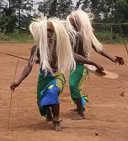 Photograph depicting two male dancers with straw wigs, neck garments, spears, and sticks
