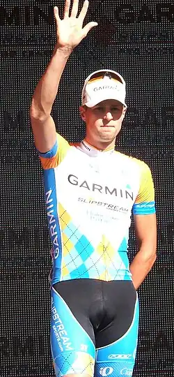 A man of about 30 wearing a gold, blue, and white argyle print cycling jersey, with his right hand in the air waving.
