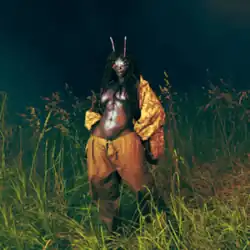 SZA standing shirtless in the middle of a grassy field with insect antennae on her head; she has yellow pants and a matching yellow jacket around her shoulders