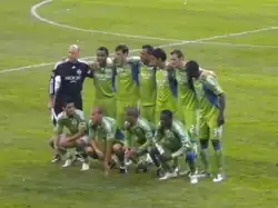 Eleven soccer players wearing green jerseys and green shorts posing in two rows on a grass field.