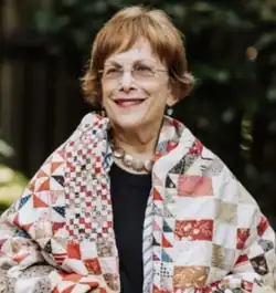 A smiling woman wrapped in a patchwork quilt. She has reddish hair worn to her ears, and wears glasses, red lipstick, dangly earrings, and a necklace of round reflective shells.