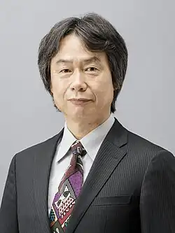 alt=Portrait photo of a Japanese man with black hair wearing a black business suit and a colorful tie.