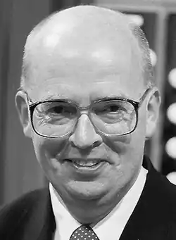 Black and white publicity headshot of a clean-shaven, bespectacled, smiling man with male pattern baldness.