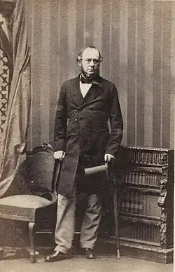 Sir John Duckworth standing between a chair to the left and low bookcase to the right, holding a top hat in his left hand