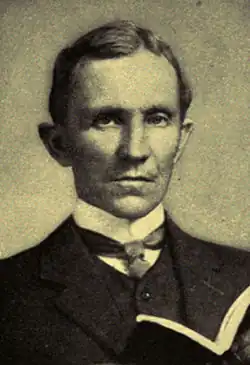 Man, probably mid-forties, dark hair, clean shaven, wearing a high collar with tie, looking straight ahead. He is holding an open book