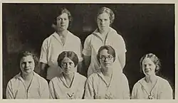 Six young women, all with mid-length hair and wearing white, early-twentieth-century athletic tops.