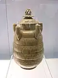 A tan jar with several rounded bands carved into the body and five upward-facing tubes equidistantly spaced along the top area of the jar. The lid is crowned with a lotus flower carving.