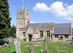 Yellow stone building, with porch with triangular roof in front. Short square tower with battlements topped by flag and flag pole. Gray gravestones in the foreground