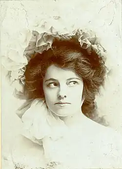 Professional monochromatic bust photograph of a white woman in an ornate white outfit; she has dark hair and is facing the camera while looking to her left.