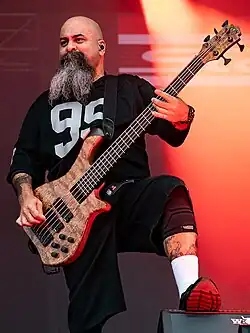 Tony Campos wearing a black print t-shirt and black shorts, playing a five-string bass guitar onstage, looking toward the audience