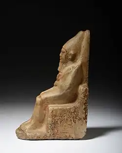 Light brown statuette of a man seated on a throne with high rounded crown, hieroglyphs inscribe on his right on side
