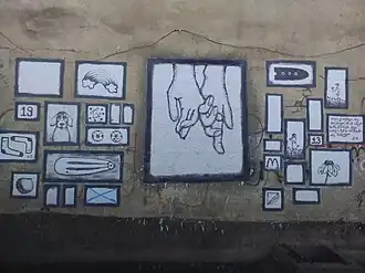 Street art on a beige wall. A central image shows two hands clasped together, surrounded by a collage of smaller, framed, black-and-white drawings of various objects and symbols.