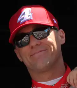 A man in his early forties smiling and waving to the crowd. He is wearing sunglasses along with a red baseball car and similarly colored racing overalls.