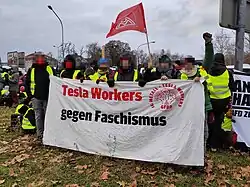 At an outdoor demonstration, six Tesla workers are carrying a banner that says "Tesla Workers gegen Faschismus" ("Tesla workers against fascism"). There is an IG Metall flag in background, and also on the banner itself. The faces of people holding the banner are blurred.