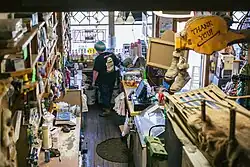 "Behind the counter with the cashier at Nagley's general store. It's an old and well-used building with creaky wooden floors and eroded countertops. There are bottles for flavored coffee, various boxes of hardware, a scale and price sticker wand, snacks, an antique cash register near a computer, an old dog sled, a pair of moccasins hanging from a sign that says Thank You, and hats that say Alaska. The front door is open.""