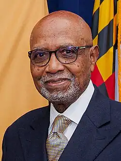 Headshot of Knox smiling in front of the flag of Maryland and a yellow cloth, wearing a dark blue suit, white shirt, and gold and blue checkered tie.