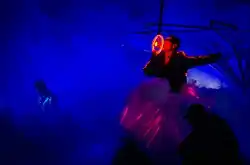 A man wearing a jacket with lights embedded in the sleeves and side, and is singing into a microphone with an illuminated circular structure around it, like a steering wheel, that he is holding with one hand.