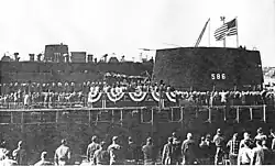 A large number of people are gathered around a platform erected just in front of a submarine.