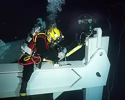 A diver in lightweight helmet and hot-water suit sits on a spreader bar while making adjustments. The emergency gas supply cylinder and excursion umbilical are clearly visible.