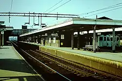 Overhead electric railway tracks with adjacent platforms in the foreground. In the background London Underground trains sit at platforms.