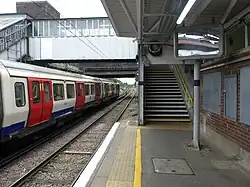Station platform with a covered footbridge connection and stairs to platform. A London Underground train is at an adjacent platform.