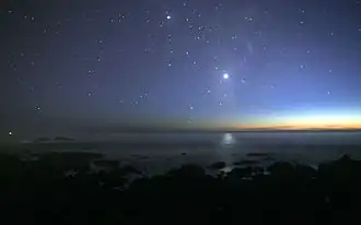 A photograph of the night sky taken from the seashore. Many glimmers of sunlight is on the horizon. There are many stars visible. Venus is at the center, much brighter than any of the stars, and its light can be seen reflected in the ocean.