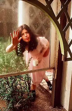blurry image of Victoria Williams wearing a pink dress with white tights underneath, black boots, and a maroon shoulder bag, standing outside a window with right hand pressed to the pane, holding a white mug in left hand, looking directly at camera with pursed lips