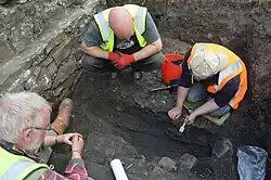 Three people crouch in a trench in the process of excavating an archaeological feature.