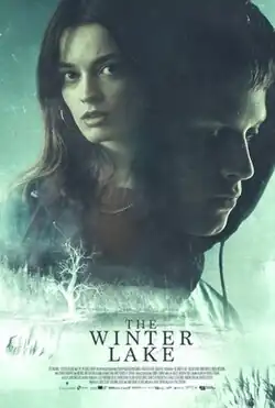 Eerie poster for The Winter Lake, featuring a young woman with a tense expression in the background and a hooded young man looking down in the foreground. Below them is a misty, desolate landscape with a barren tree and a frozen lake. The title The Winter Lake is displayed prominently at the bottom in bold, icy lettering.