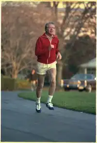 Color photo of Carter in a red jacket and white shorts jogging on a road