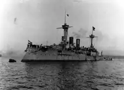 A large warship moored to a buoy, with a group of sailors crowded together on the bow