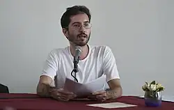 Un hombre joven visto de frente, detrás de una mesa hablando al micrófono, usando barba y lentes.