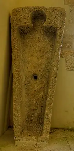 Photo couleur d'un bloc de pierre placé verticalement. Il est creusé pour accueillir une dépouille funéraire. Une loge plus petite est découpée pour recevoir le cou et la tête.