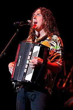 Una fotografía de "Weird Al" Yankovic, cantando a través de un micrófono y tocando una armónica.