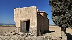Ruinas de la iglesia con un pino a un lado, el resto del paisaje es todo un bancal de tierra sin plantar. Se aprecia que la puerta está abierta y hay una pequeña valla para evitar el acceso.