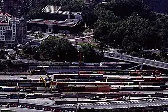 Estación de tren con muchísimos contenedores de carga, un tren de cercanías y la ría detrás