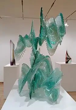 Vista lateral de la pieza Brote (1992). Escultura de vidrio laminado transparentes con varias capas pegadas unas con otras, creando una figura que parece dinámica. Al fondo de la sala se observan otras dos esculturas, estas de colores