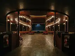 Vista de la sala del teatro Margarita Xirgu desde la entrada a la misma. PIso de madera y pocas butacas rojas. De fondo, en el escenario, un telón azul.
