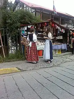 Dos mujeres colonieras, vestidas con la indumentaria tradicional de la Colonia Tovar, frente a un puesto de comida y productos típicos de la región.