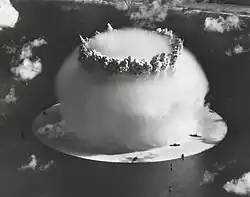 Aerial photo of nuclear explosion rising from lagoon. Hemispherical condensation cloud on the surface is 1 mile (1.6 km) in diameter. In comparison, Navy ships in the foreground look like bathtub toys.
