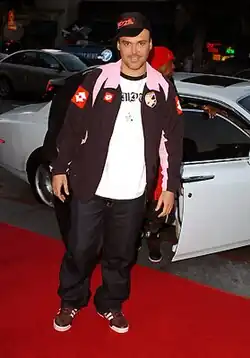 A man standing before a car. He is wearing a white T-shirt and brown coat, with black pants and sneakers