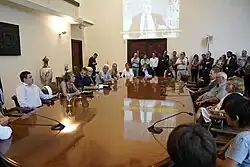 Imagen de la sala de Acuerdos de la intendencia de Montevideo con gran cantidad de personas presentes y al fondo una proyección de imagen de Julio Baráibar.
