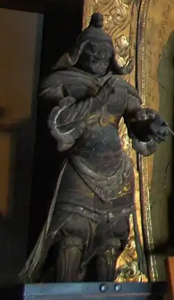 Three-quarter view of a standing statue. The right arm is bend with the right hand in front of the breast. The left arm is also bend with the palm of the left hand pointing upward.