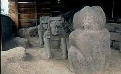 Tres esculturas olmecas de basalto dentro de una estructura que protege un sitio arqueológico. En primer plano a la derecha de espaldas se observa la figura de un jaguar, frente a ella dos esculturas similares de hombres hincados sosteniendo un bastón, ataviados con un taparrabo y un tocado complejo