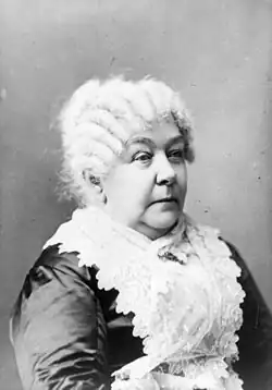 A monochrome photograph portrait of an elderly woman shown from the elbow up, turned somewhat to the right, a white lace apron covering a dark shiny fabric dress, the white hair in front-to-back rows of ringlets